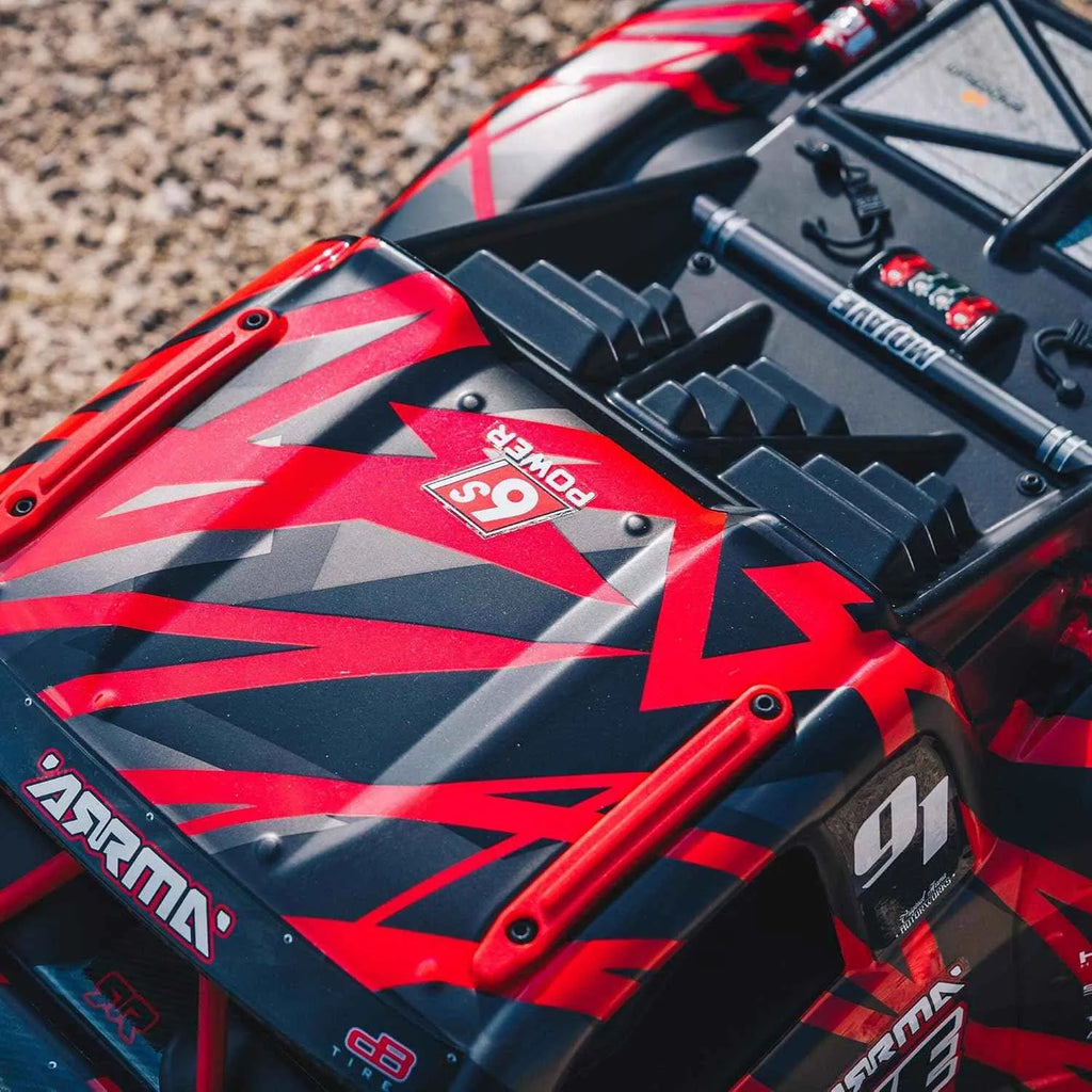 Close-up of a red and black RC short course truck with visible branding on a gravel surface.