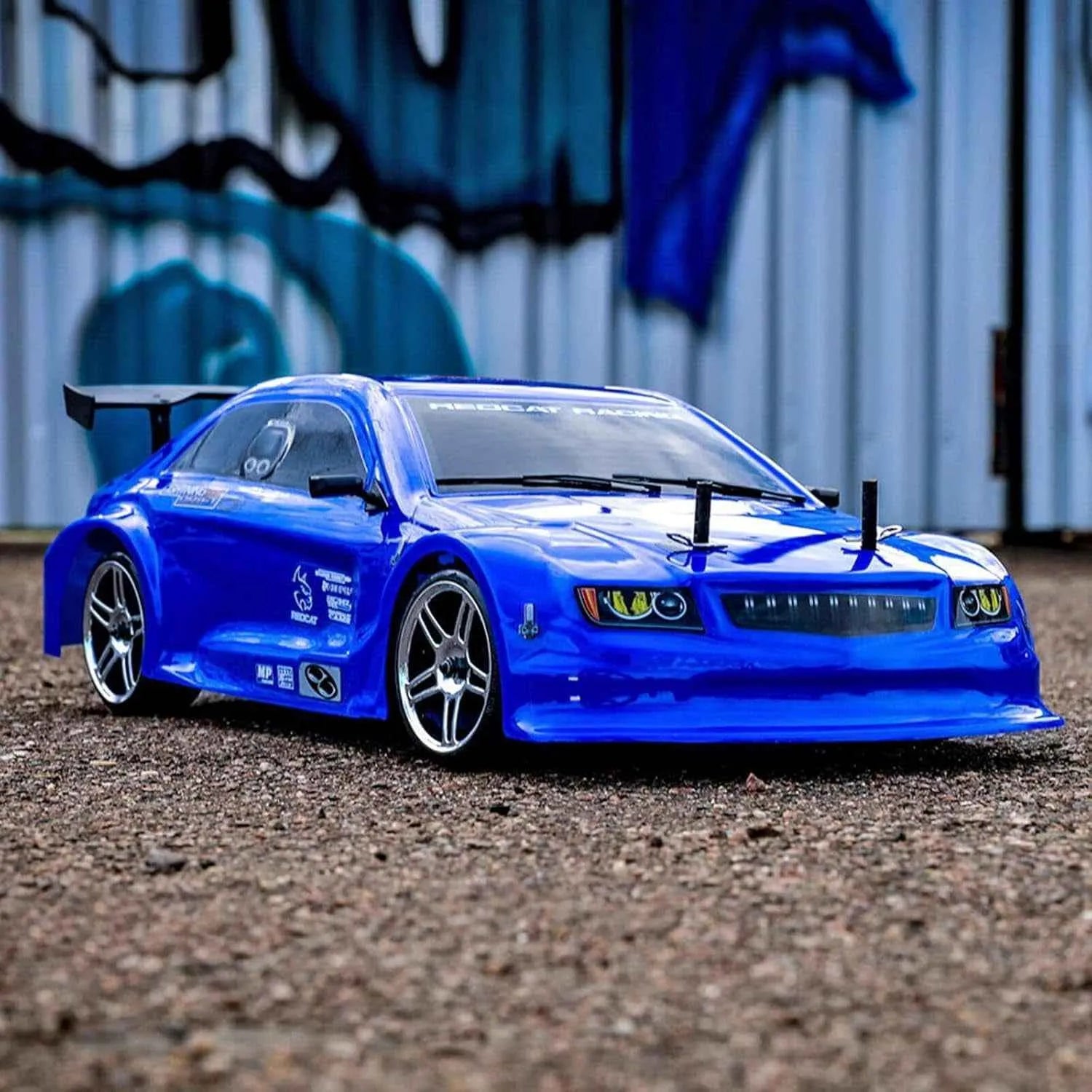 Blue rc drift car on a gravel surface with a graffiti-covered wall in the background
