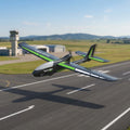 Green and gray RC plane on a runway with airport and mountains in the background